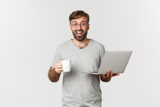 Young happy man working with laptop, drinking coffee and smiling, standing over white background.