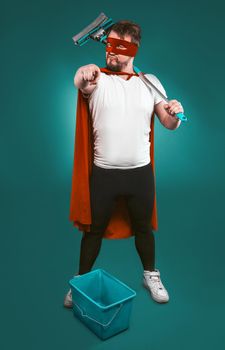 Superhero is ready to fight for cleanliness, Brave man in a red superhero uniform with a bucket and a mop points the camera with his index finger. Cleaning concept. Isolated on green.