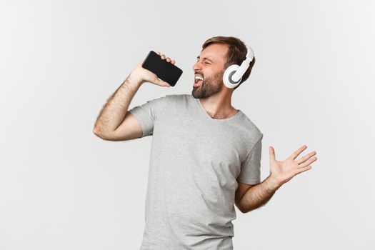 Portrait of handsome guy in gray t-shirt, playing karaoke app in headphones, singing in mobile phone microphone, standing over white background.