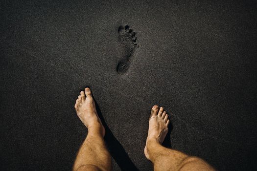 Close up of male footprints and feet walking on the volcanic black sand on the beach.