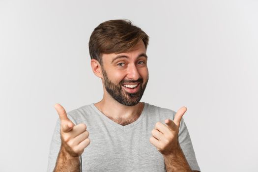 Close-up of handsome adult man with beard, smiling and pointing fingers at camera, standing in gray t-shirt over white background.