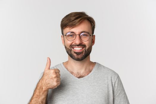 Close-up of cheerful caucasian man with beard, wearing glasses and casual t-shirt, smiling and showing thumbs-up in approval, recommend product, standing over white background.