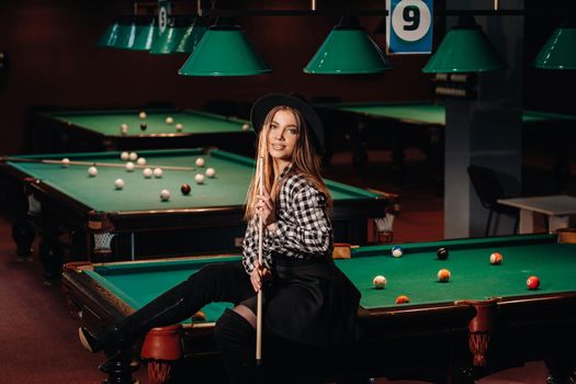 A girl in a hat in a billiard club sits on a billiard table with a cue in her hands.Playing billiards.