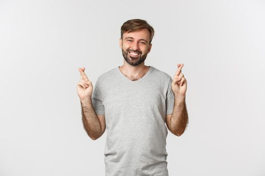 Portrait of optimistic handsome man in gray t-shirt, hoping for something with fingers crossed, making wish and smiling, standing over white background.
