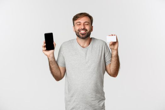 Handsome young man shopping online, holding credit card and mobile phone, showing smartphone screen, standing over white background.