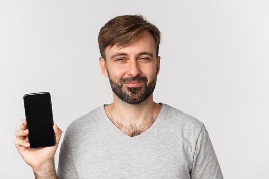 Close-up of pleased smiling man with beard, looking delighted and showing mobile phone screen, standing over white background.