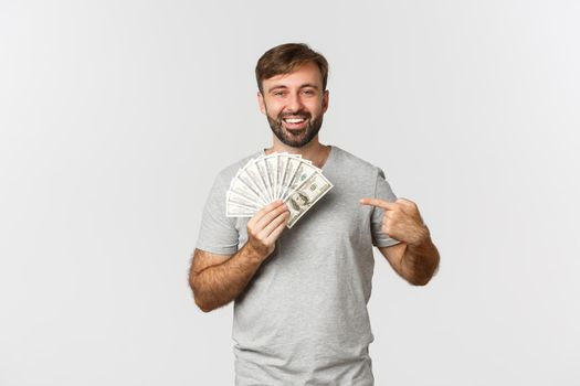 Portrait of handsome bearded male model in gray t-shirt, pointing at money and smiling, getting a loan, standing over white background.