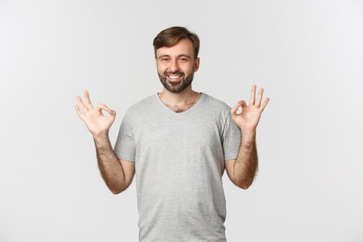 Portrait of handsome smiling man in casual grey t-shirt, showing okay sign in approval, agree to you, praising good choice, standing over white background.