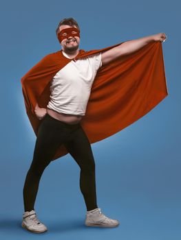 Smiling super hero man ready to fly save world. Funny man in red superhero costume and mask holding his cloak with hand posing in full growth on faded denim blue background.