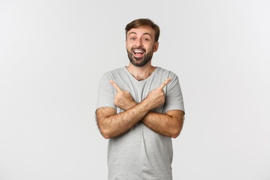 Portrait of happy and amazed bearded male model in gray t-shirt, pointing fingers sideways, showing two good choices, standing over white background.