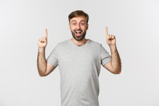 Cheerful handsome man in gray t-shirt, showing logo or advertisement, pointing fingers up and smiling happy, standing over white background.