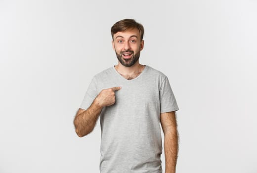 Image of happy and surprised man in gray t-shirt, pointing at himself, standing over white background.