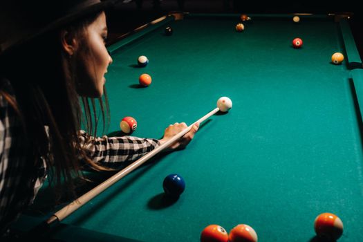 A girl in a hat in a billiard club with a cue in her hands hits a ball.Playing billiards.