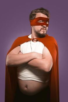 Super hero man with crossed arms and opened tummy looking at camera. Serious man in cape and superhero costume isolated on grape purple background. Power concept.