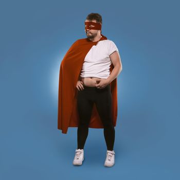 Body positive Super hero man added weight over winter. Sad man in superhero costume checks fat on his tummy. Isolated on blue background. Template with place for text. Overweight concept.