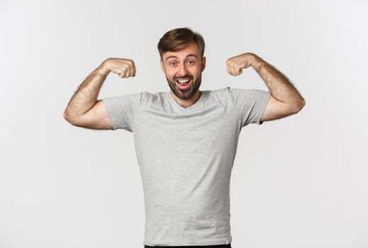 Portrait of confident bearded man flexing biceps, showing his muscles after gym workout, standing over white background.