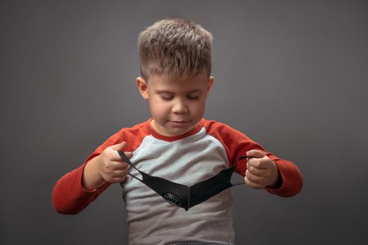 Preschool boy holds protective face mask looking on it. Quarantine concept.