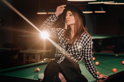 A girl in a hat in a billiard club sits on a billiard table.Playing pool.