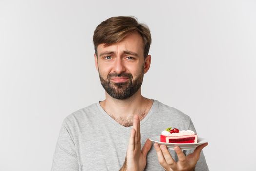 Close-up of reluctant bearded man, declining piece of cake and grimacing, standing over white background.