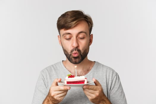 Close-up of happy bearded man, smiling and celebrating birthday, holding cake with lit candle, making b-day wish, standing over white background.