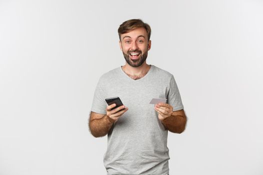 Handsome young man shopping online, holding credit card and mobile phone, standing over white background.