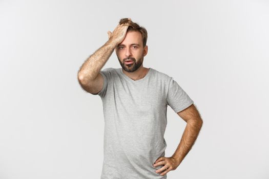 Troubled bearded man in gray t-shirt, slap forehead and sighing bothered by problem, standing over white background distressed.
