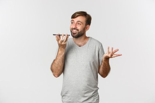 Image of carefree guy with beard, wearing basic t-shirt, talking on speakerphone, holding mobile phone near mouth to record voice message, standing over white background.