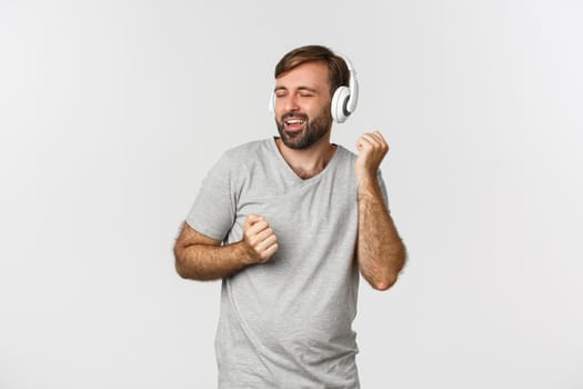 Portrait of happy handsome guy in gray t-shirt, listening music in wireless headphones and dancing, standing over white background.