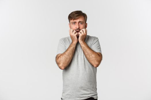 Portrait of scared caucasian guy in gray t-shirt, holding hands near mouth and trembling from fear, standing frightened over white background.