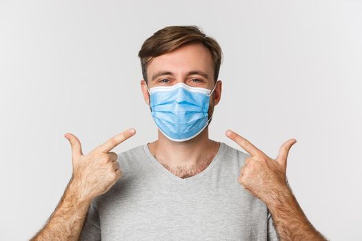 Concept of pandemic, covid-19 and social-distancing. Close-up of cheerful man recommend wear medical mask during coronavirus, standing over white background.