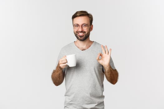 Cheerful young man with beard, wearing glasses and gray t-shirt, drinking coffee and showing okay sign in approval, recommend something, standing over white background.