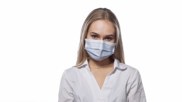 Looks sullenly young nurse in a medical mask and white uniform with blond straight hair looking at the camera. Isolated on white background.