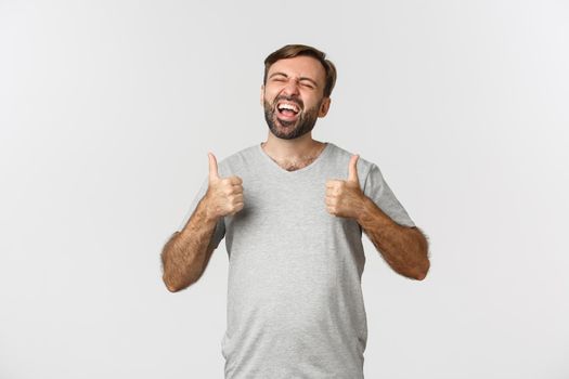 Portrait of happy and excited guy with beard, laughing over something funny and showing thumbs-up, standing over white background.