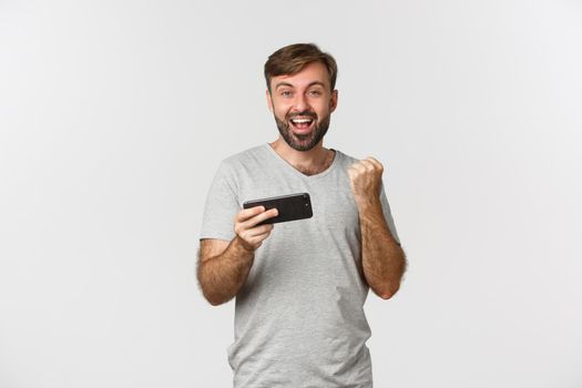 Portrait of happy man in gray t-shirt, holding smartphone and rejoicing, triumphing over achievement, standing over white background.