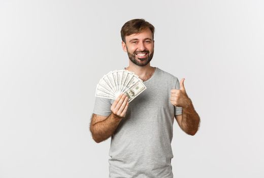 Portrait of handsome man with beard, showing thumbs-up and holding money, recommend credit or loan, standing over white background.