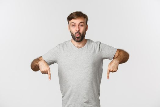 Portrait of excited guy with beard, pointing fingers down at something curious, showing logo, standing over white background.