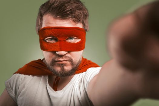 Super hero man makes selfie photos or video by mobile phones. Serious man wearing red mask and cloak posing for his social networks followers. Isolated on green background.