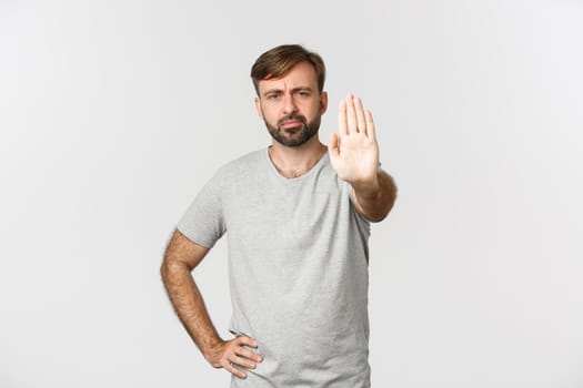 Portrait of serious frowning guy in gray t-shirt, showing stop gesture, disapprove and prohibit action, standing over white background.