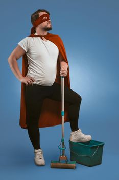 Super cleaning from superhero. Brave guy ready to clean up and fight dark forces with help of cleaning equipment. Caucasian man looks forward holding mop with his foot on bucket on blue background.