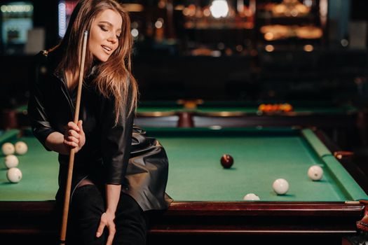 A girl with a cue in her hands sits on a table in a billiard club.Russian billiards.