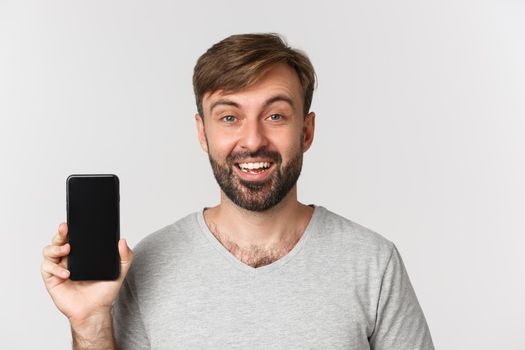 Close-up of smiling handsome man in gray t-shirt, showing mobile phone screen, recommending app or shopping site, standing over white background.