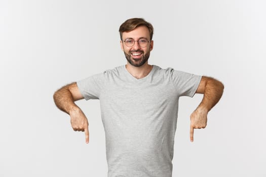 Cheerful bearded man smiling, pointing fingers down, showing logo, wearing gray t-shirt, wearing gray t-shirt, standing over white background.