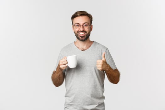 Cheerful young man with beard, wearing glasses and gray t-shirt, drinking coffee and showing thumbs-up in approval, recommend something, standing over white background.