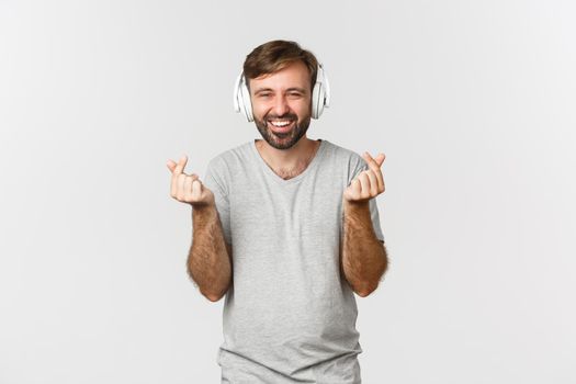 Image of happy caucasian man in gray t-shirt, listening music in headphones and showing heart signs, smiling at camera, standing over white background.