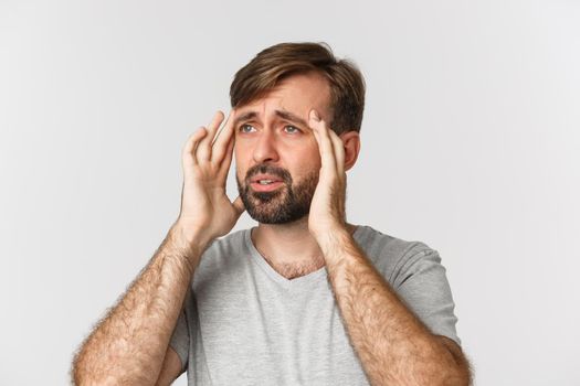Close-up of gloomy bearded man, touching head and grimacing, having a headache or migraine, standing over white background.