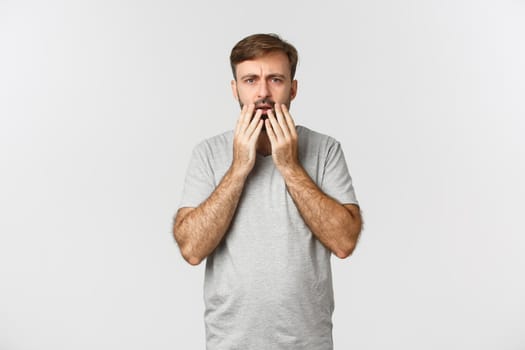 Portrait of shocked and worried guy staring at something disturbing, gasping and frowning, standing over white background.