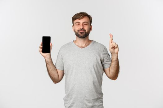 Portrait of hopeful man in gray t-shirt, showing mobile phone screen and making wish, crossing fingers for good luck, standing over white background.