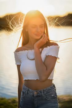 Happy teenage girl smiling with backlit sunlight at sunset in nature. Charming Caucasian young lady on background of river. Summer vacation concept.