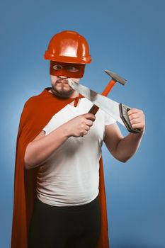 Superhero builder or repairman holds construction tools. Man in red helmet and Superhero costume crossed saw and hammer while looking at camera while standing on a denim blue background.