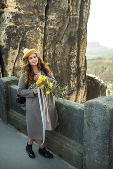 a girl in a gray coat and hat with a bouquet of flowers on the background of mountains and gorges in Swiss Saxony, Germany, Bastei.